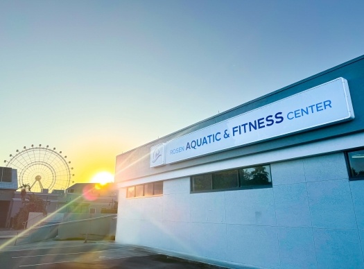 A building with a sign that reads “Rosen Aquatic and Fitness Center” at sunset, with a sun glare and a Ferris wheel in the background. We hope you enjoyed this photo tour of the Rosen Aquatic and Fitness Center.