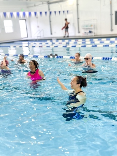Six people in chest-deep water in an indoor pool with blue and white lane markers in the background and a string of blue and white flags overhead. They are participating in an aquatic fitness class at Rosen Aquatic and Fitness Center.