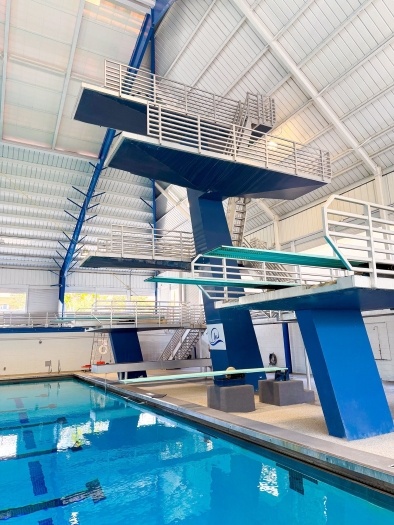 Three large diving platforms with railings against a white metal ceiling with the corner of a blue pool below. 
