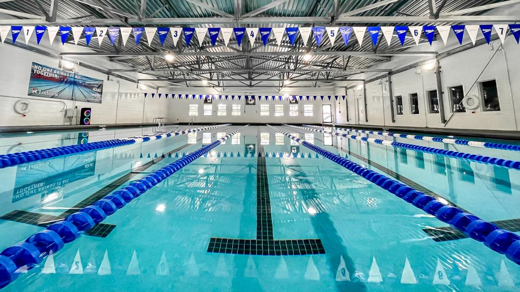 Bright indoor swimming pool with turquoise water, blue and white lane dividers, and blue and white numbered flags overhead.