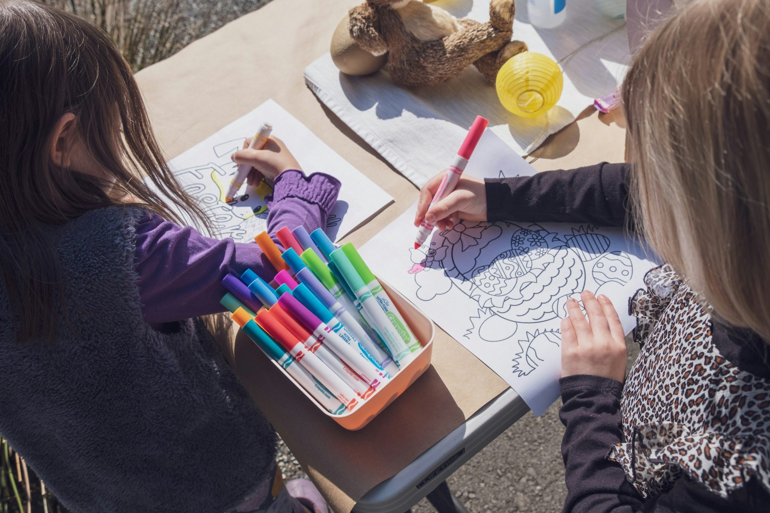 Two kids are coloring on white sheets of printed paper at a light beige table outside with a box full of colorful markers, a teddy bear, and a yellow ball also on the table.