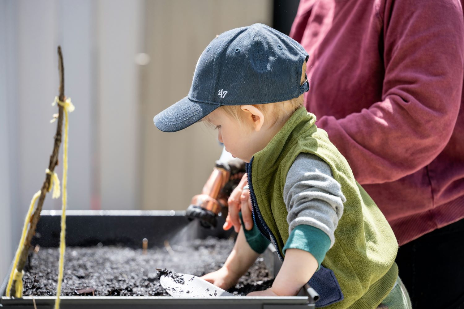 A young child in a blue cap and green vest digs in a black planter with dark soil using a white trowel, while an adult in a maroon shirt watches nearby outdoors. 