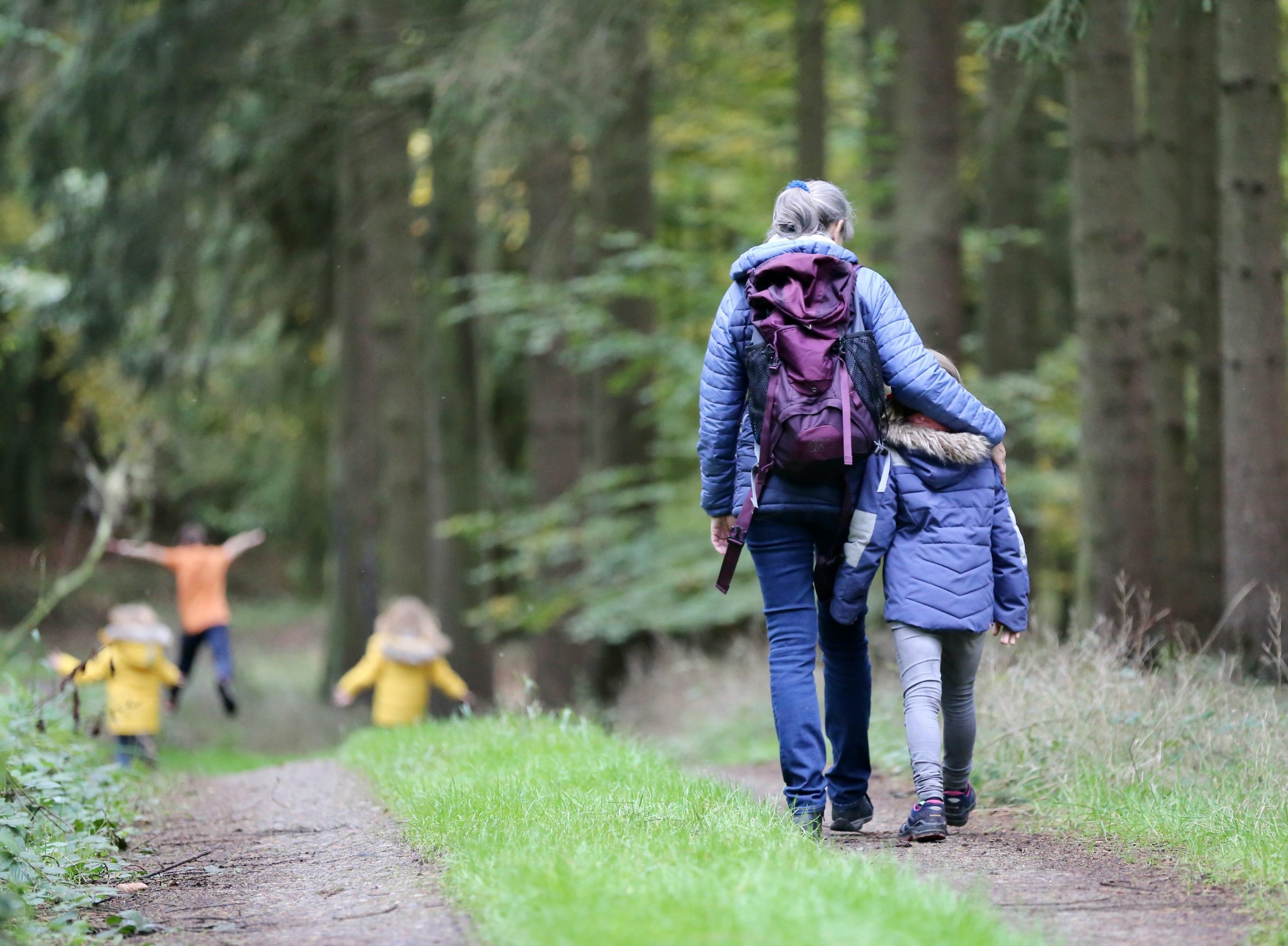A woman in jeans and a blue jacket puts her arm around a child with a blue jacket and jeans as they walk surrounded by trees and a dirt trail, with two kids in yellow jackets in the distance. Walking is a great healthy activity to do with kids.