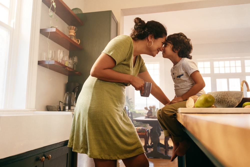 A mom holding a coffee mug and her toddler son lean in forehead to forehead in the kitchen. 