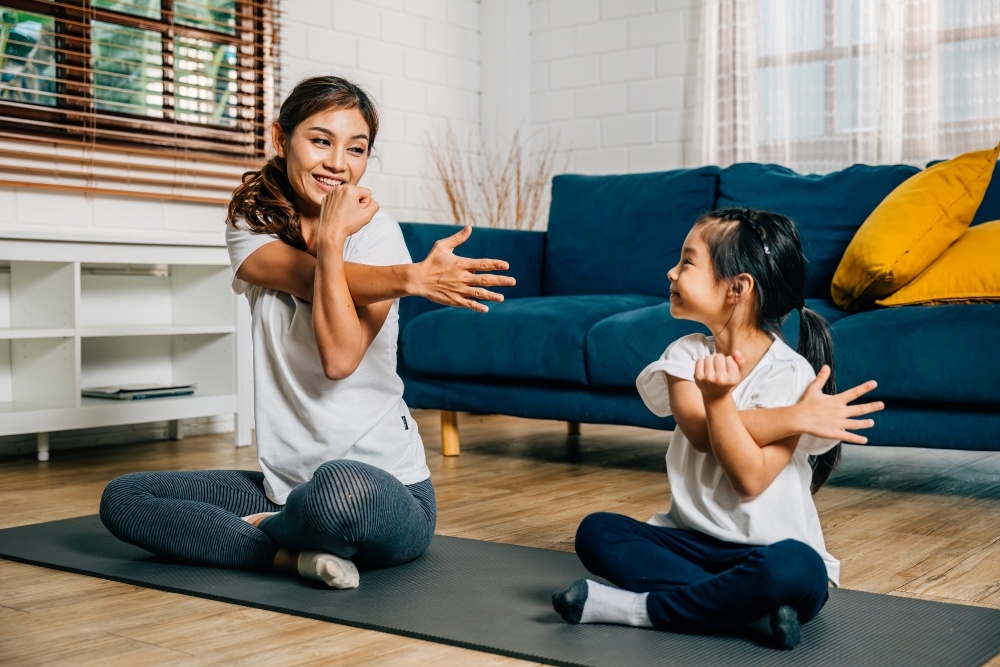A mom and her daughter sit on a yoga mat, stretching their shoulders together. 