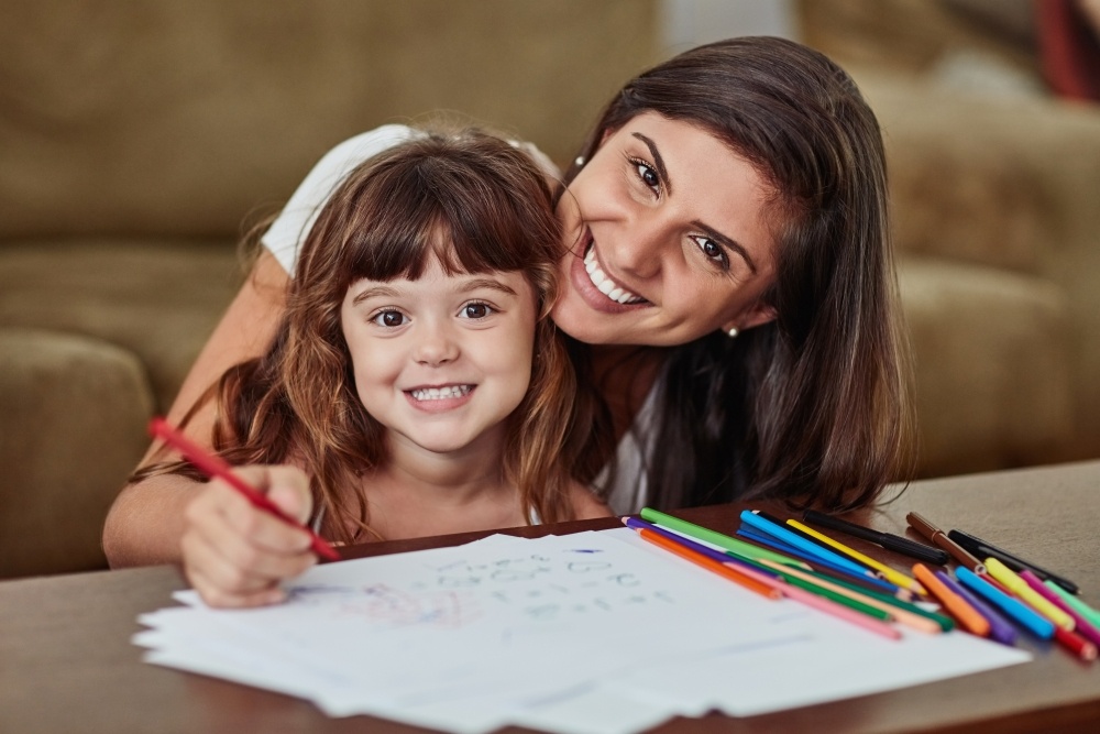 A mom and her daughter color together, smiling at the camera. 