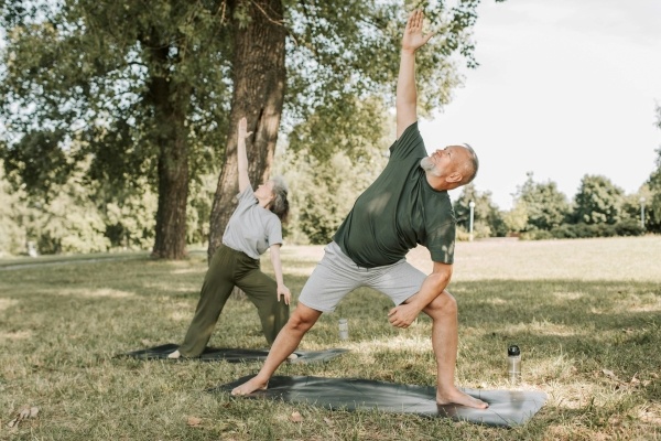 And older man and woman stand on yoga mats in a field beside trees, bent over and stretching up to the sky.