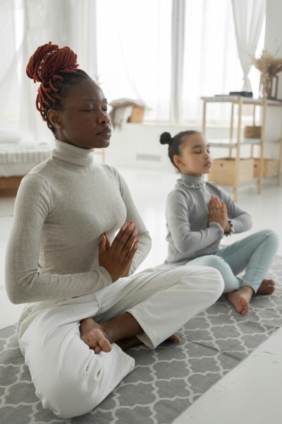 A woman and young girl in comfortable clothes sit cross-legged on a yoga mat with eyes closed and their hands folded at their hearts.