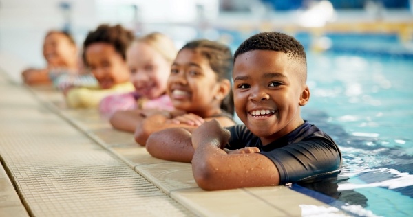 A row of five children with their arms crossed leaning on the side of an indoor swimming pool, smiling. 