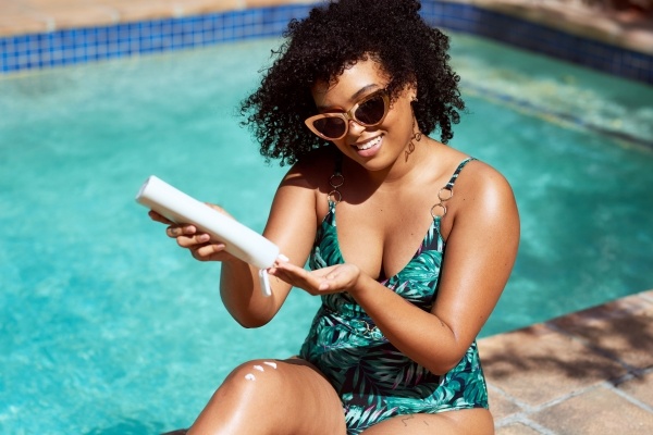 A woman in sunglasses and a green floral bathing suit sits by a pool and squeezes sunscreen into her hand.
