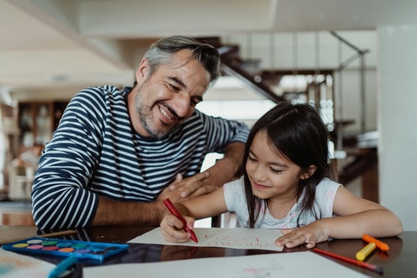 A dad and his daughter create a sign with crayons and paper at a table together.