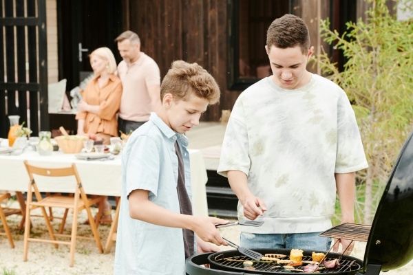 Two teen boys flip food on a round grill while an older couple embraces in front of a set table in the background.