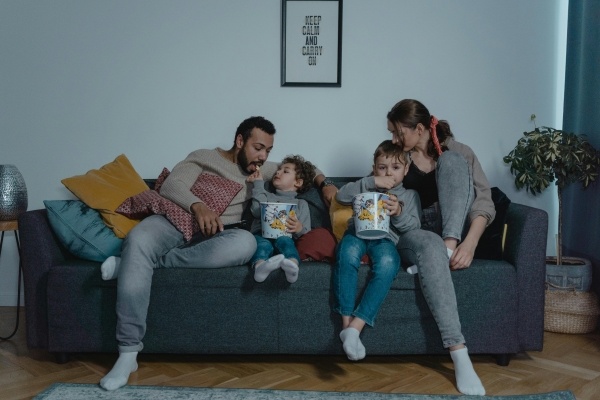 A mom and dad with two kids holding popcorn buckets between them sit on a sofa in a dim room.