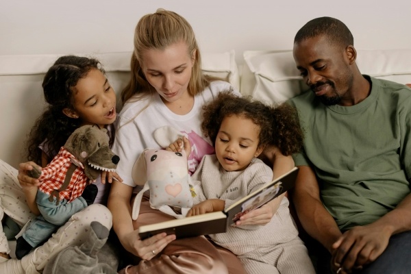 A mom and dad sit on a couch with two young girls reading a book together.