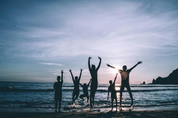 A family of six people silhouetted jumping in the shallow waves of the ocean against a blue sunset sky. Go to the beach to celebrate Family Fun Month.