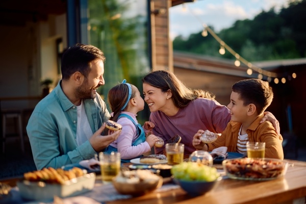 A mom, dad, and two young kids sit at an outdoor restaurant table smiling while eating donuts and surrounded by plates of food.