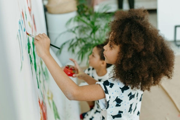 Two young girls stand before a large white banner painting with paintbrushes and a jar of red paint.