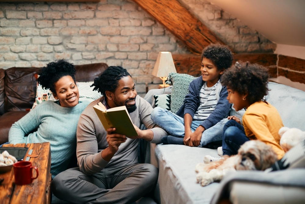 A mom and dad sit on the floor reading a book to two children on a couch beside them during Family Fun Month. 