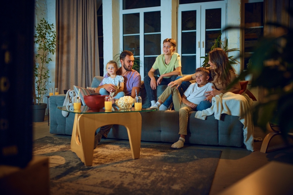 A mom, dad, and three kids sit on a couch in a cozy and dimly lit living room for family movie night. 