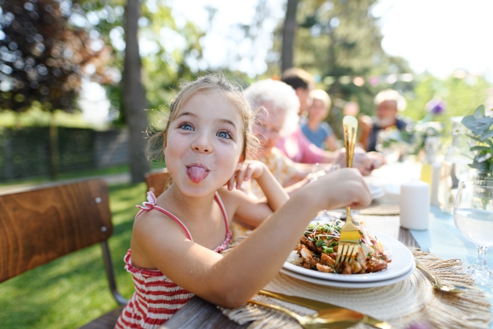 A young girl sits at an outdoor table with other people sticking her tongue out at the camera playfully. 