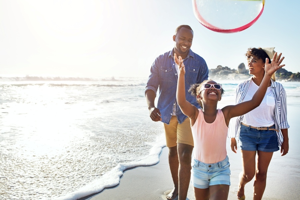 A mom, dad, and little girl walk along the shore of a sunny beach, while the girl throws a beach ball in the air. 
