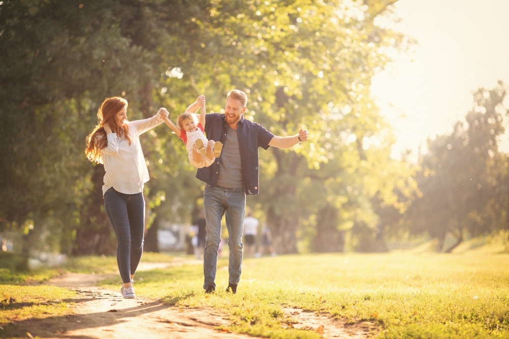 A mom and dad walk on a trail outdoors, swinging their toddler by the arms between them during Family Fun Month.