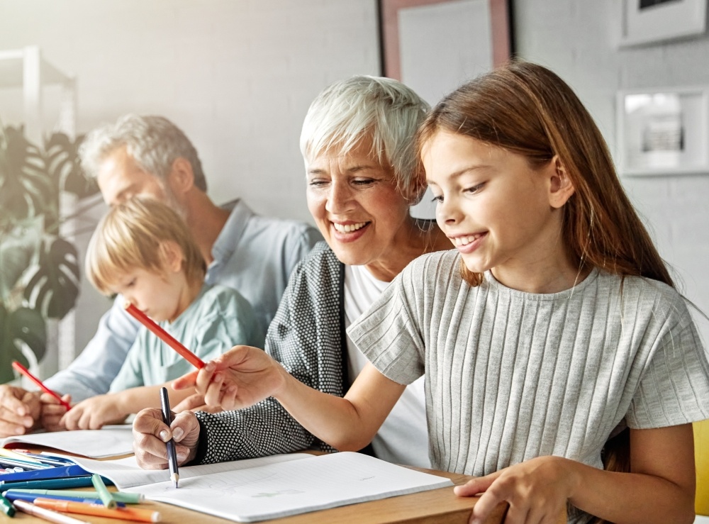 A grandmother and grandfather sit at a table with their two grandchildren writing in a notebook.