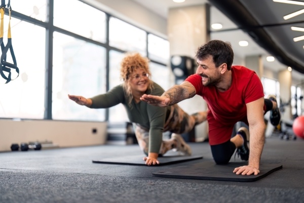 A man and woman each kneeling on a yoga mat with an arm outstretched, smiling at one another.