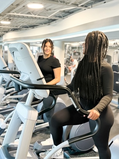 Two women in black workout gear smile at each other while on elliptical machines.
