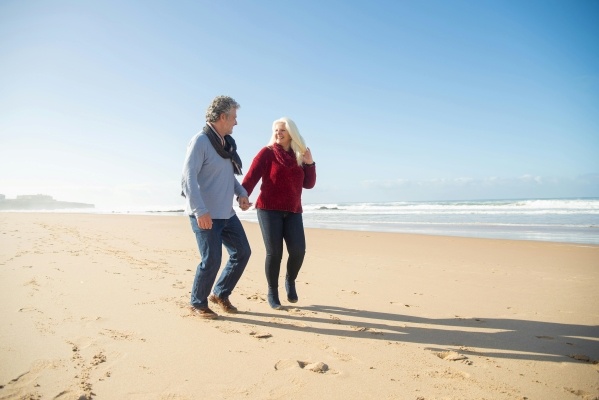 An older couple holding hands while walking on a beach in the warm sun.