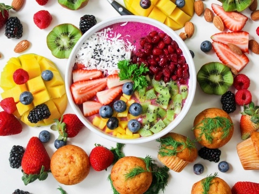 An overhead shot of a display of muffins, fresh fruit like strawberries and mangos, and an acai bowl with pomegranate seeds, kiwi, berries, and coconut.