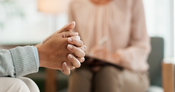 A close-up of a woman's clasped hands resting on her lap with a woman taking notes out of focus in the background.