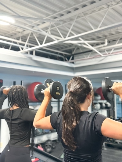 The backs of two women in black work out clothing as they lift dumbbells.