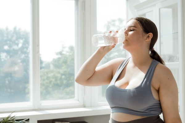 A woman in a gray sports bra drinks a glass of water in front of bright windows.