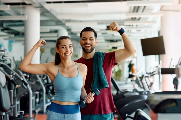 A man and a woman in workout gear flex their arms in an indoor gym.