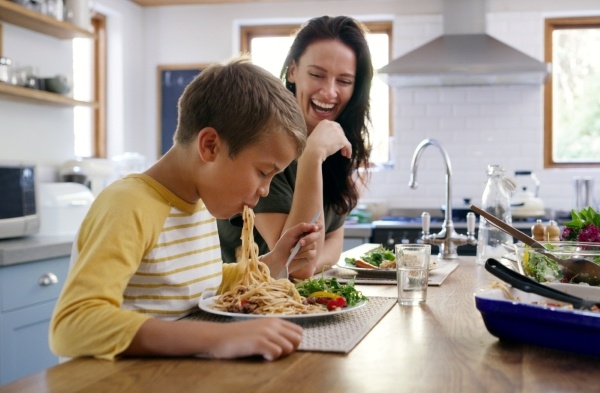 A boy slurps spaghetti sitting at a kitchen counter while his mom looks on and laughs during family dinner.