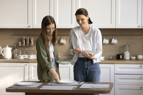 A mom and her teenage daughter set the kitchen table for a family meal together.