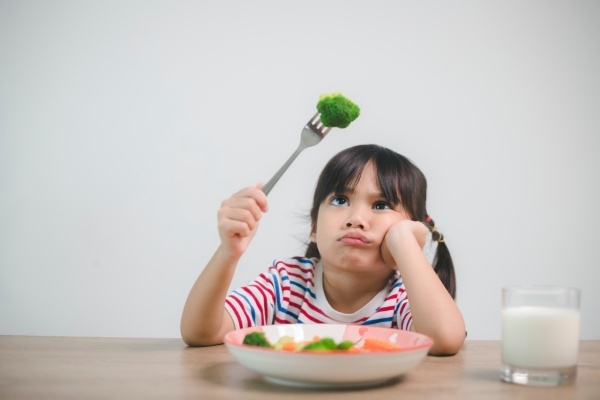 A young girl sits at a table with a plate of food, staring unhappily at a piece of broccoli on her fork.