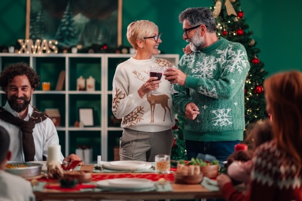 And older couple in Christmas sweaters stand and toast with wine glasses in a room decorated for Christmas and surrounded by happy people.