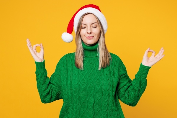 A woman in a green sweater and Santa hat stands with meditative hands and closed eyes, practicing a healthy holiday mindset.