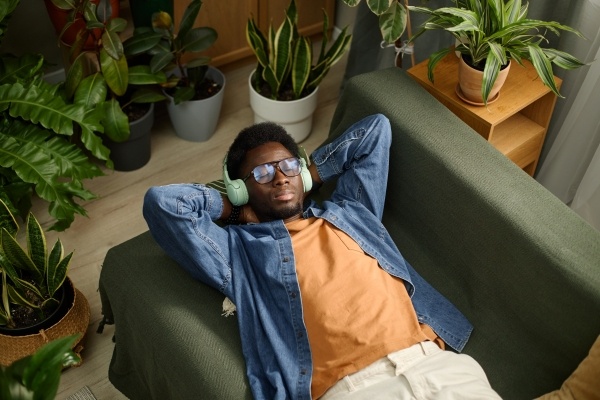 A man lays on a green couch wearing headphones and surrounded by houseplants as he practices mindfulness.