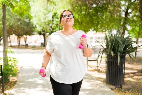 A smiling woman walking outside in workout clothes, carrying light dumbbells. 
