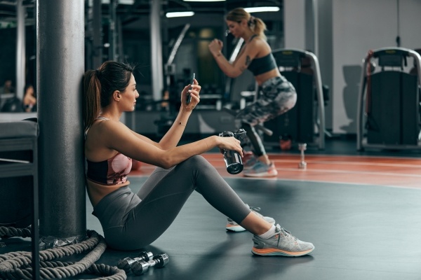A woman in workout clothes sits on the floor at a gym, scrolling through her phone.
