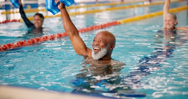 An older man lifts a water weight overhead, smiling while standing neck-deep in pool water.