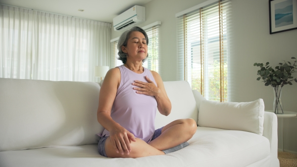 An older woman sits on a couch in a bright room with her eyes closed, legs crossed, and her hand on her chest peacefully as she focuses on mental health.