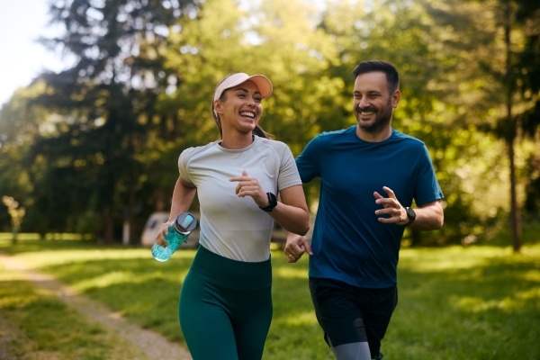 A man and a woman in athletic clothing run and laugh in a green outdoor space.