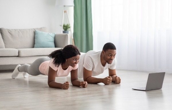 A couple in athletic clothing planks together while looking at a computer screen during a couple's workout.