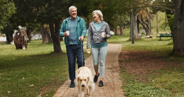 An older couple in athletic clothing walk a golden retriever outdoors while holding coffees and smiling.