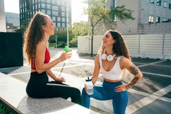 Two women in an urban setting wearing athletic clothing smile at one another while taking a water break.