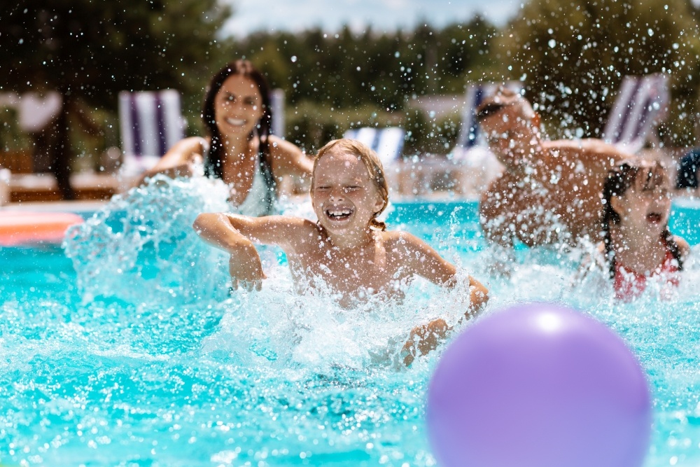 A family with two kids splash and laugh in the pool with a purple ball in the foreground.